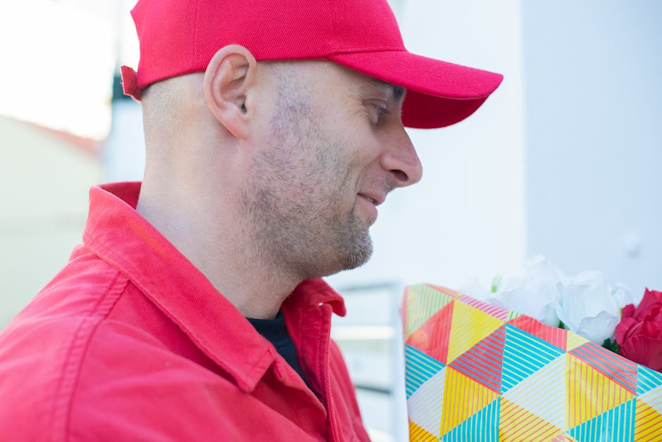 A delivery person wearing a red cap and red jacket is holding a colorful bouquet of fresh flowers wrapped in decorative paper. The bouquet contains vibrant pink roses and white blooms, arranged neatly