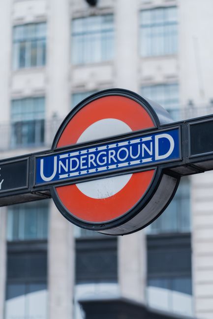 Close-up view of a London Underground sign featuring the iconic red circle and blue bar with 'UNDERGROUND' text, mounted on a black metal framework. In the background, there is a modern building with