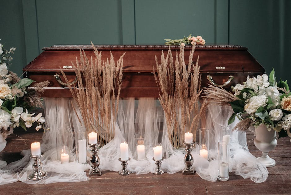 A wooden funeral casket placed on a dark wooden floor, decorated with an arrangement of white and cream-toned flowers including roses and lilies, flanked by large white vases filled with similar bloom
