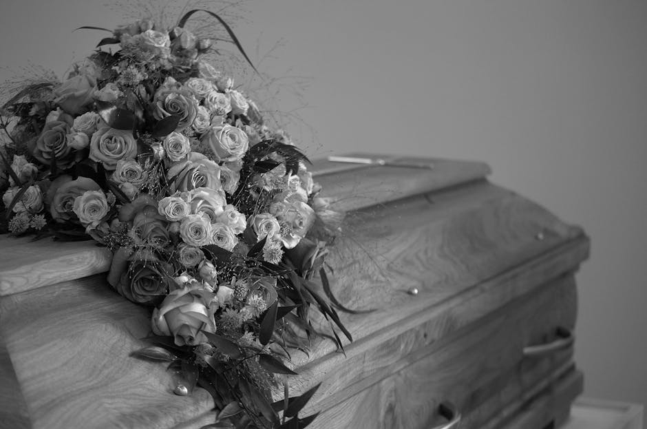 A large, elegant floral arrangement of fresh roses and other flowers, predominantly in soft, pastel shades, resting atop a wooden casket with a smooth, polished surface. The bouquet features a variety